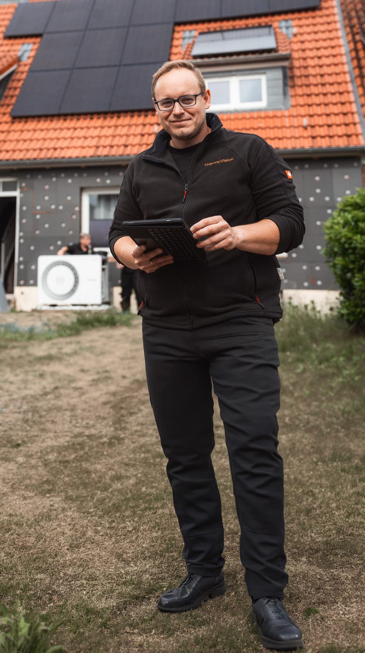 ThermoVision Mitarbeiter mit Tablet vor einem Einfamilienhaus mit Photovoltaikanlage und Wärmepumpe