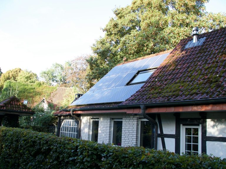 Haus mit rotem Steildach und Solarpanel, umgeben von grünen Bäumen und Sträuchern.