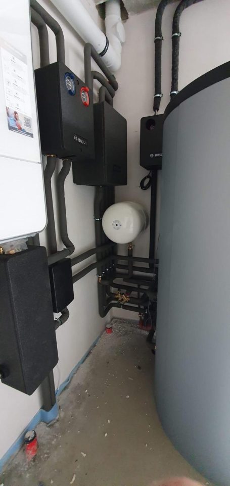 Rohrleitungen und Geräte in einem Technikraum, grauer Wassertank an der Wand.