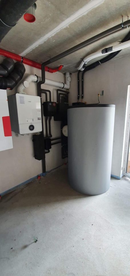 Grauer Heizkessel in einem Technikraum mit Rohren und Steuergeräten an der Wand.